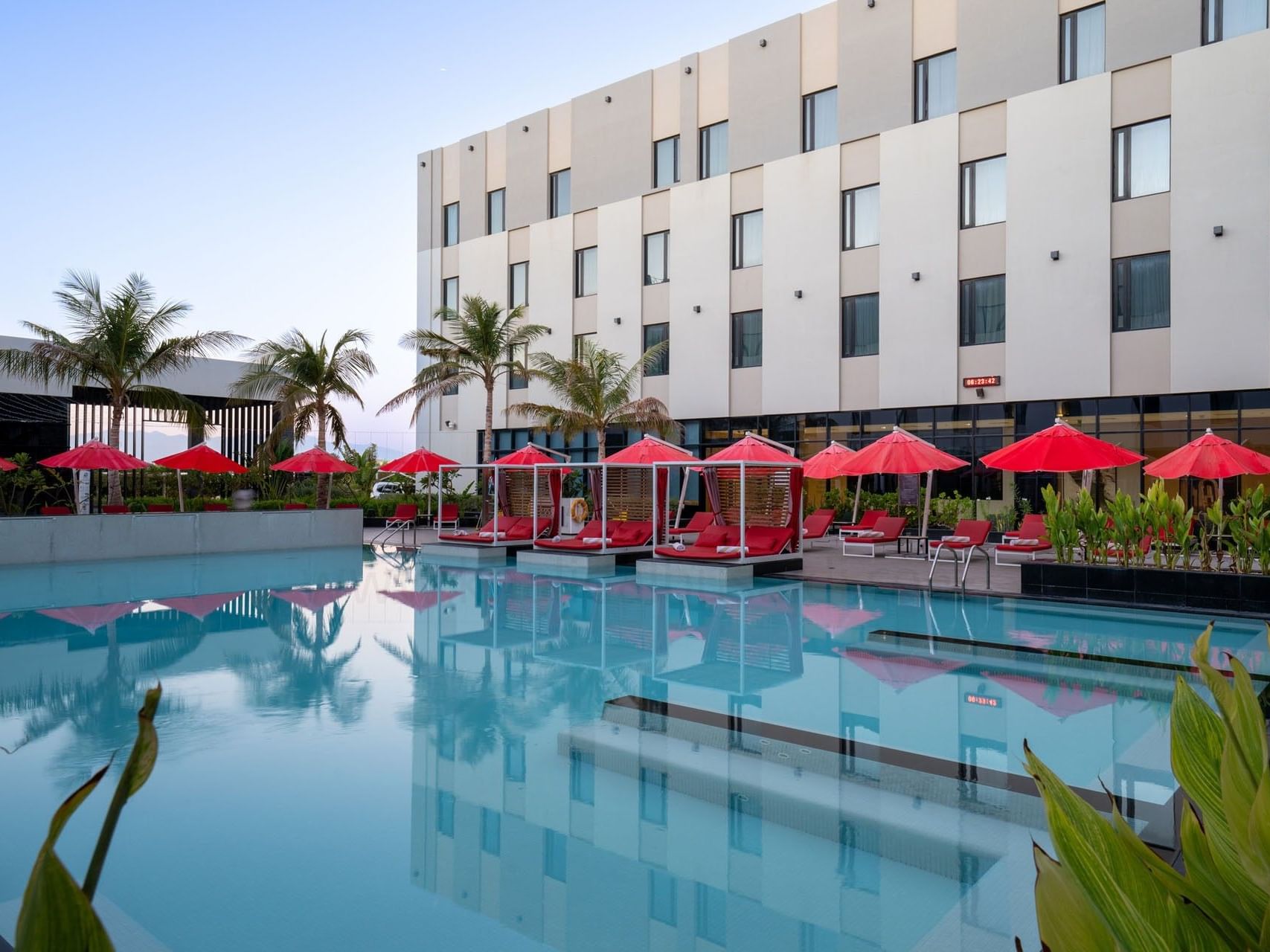 Modern hotel with outdoor pool, lounge chairs, and red umbrellas under clear sky.