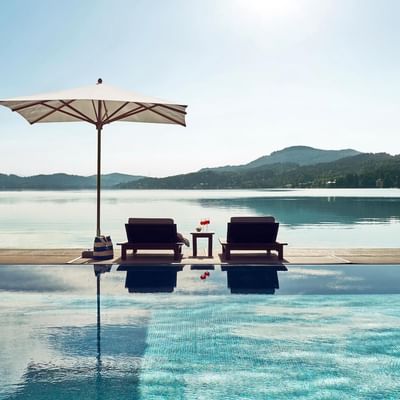 Pool mit zwei Liegen und Sonnenschirm neben einem See mit Bergblick.