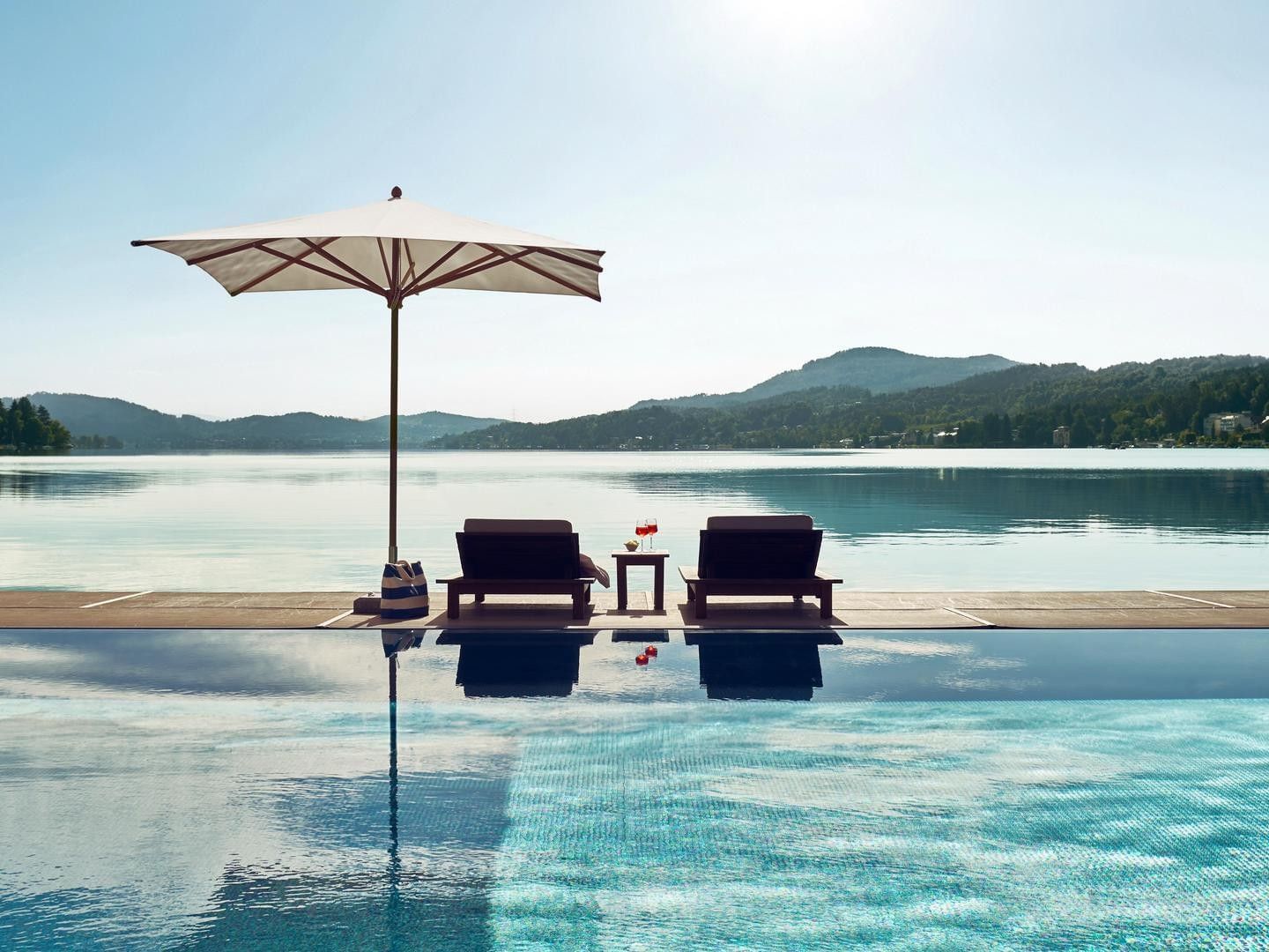 Piscina con ombrellone e lettini, vista lago e montagne sullo sfondo.