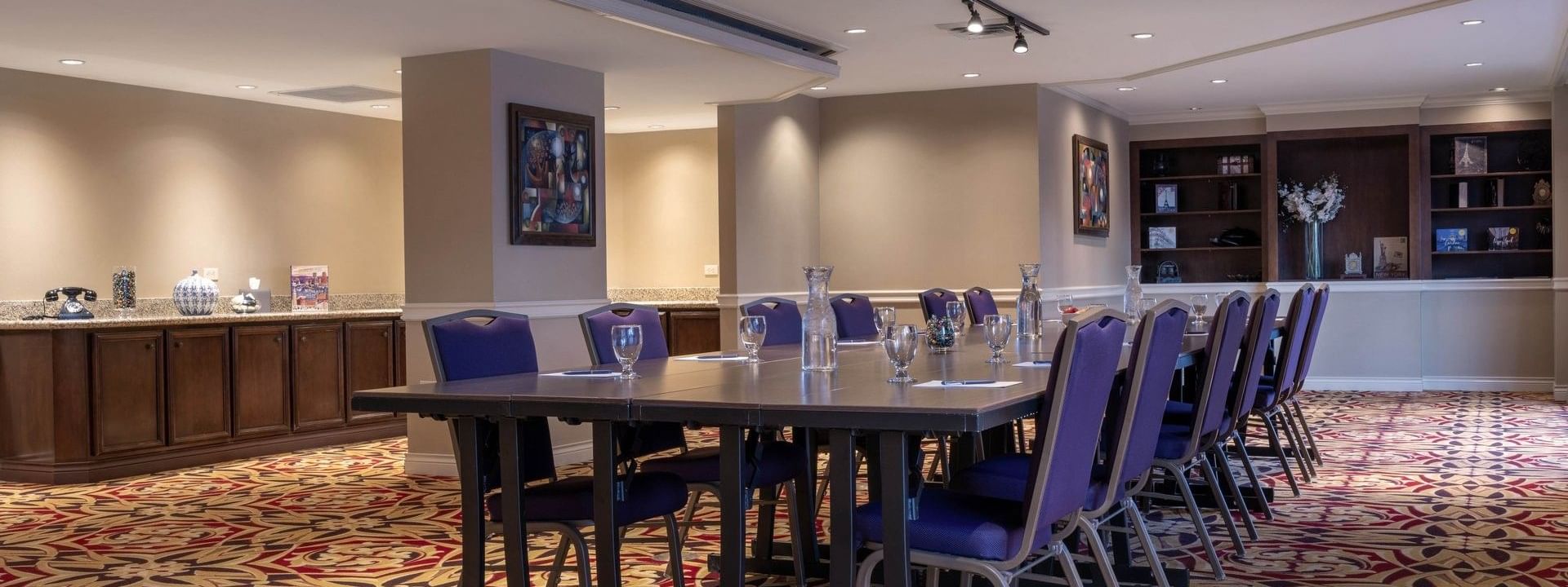 Long dark table by purple chairs under track lighting on a patterned rug in Executive Lounge at Warwick Denver