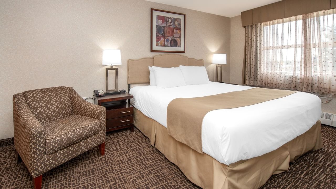 A hotel room with a bed, nightstand, lamp, chair, and a framed art on the wall.