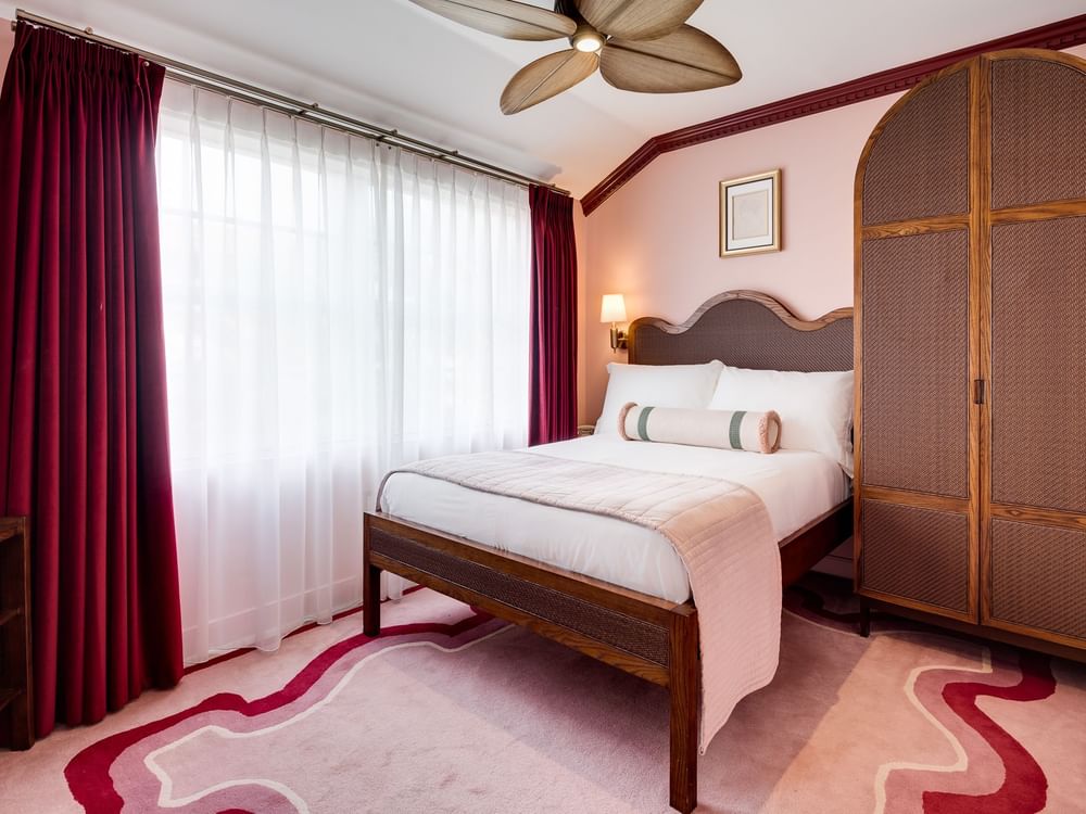 A hotel room with a king-sized bed, a large wardrobe, and a window with red curtains.