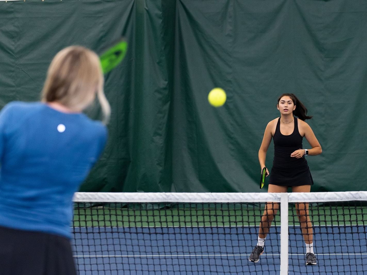 Two women play pickleball on an indoor court, one poised to hit the ball.