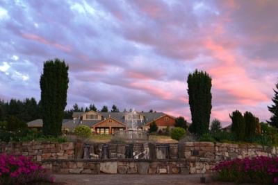 A serene outdoor scene featuring a stone wall, a fountain, flowering plants, and a house under a colorful sky.