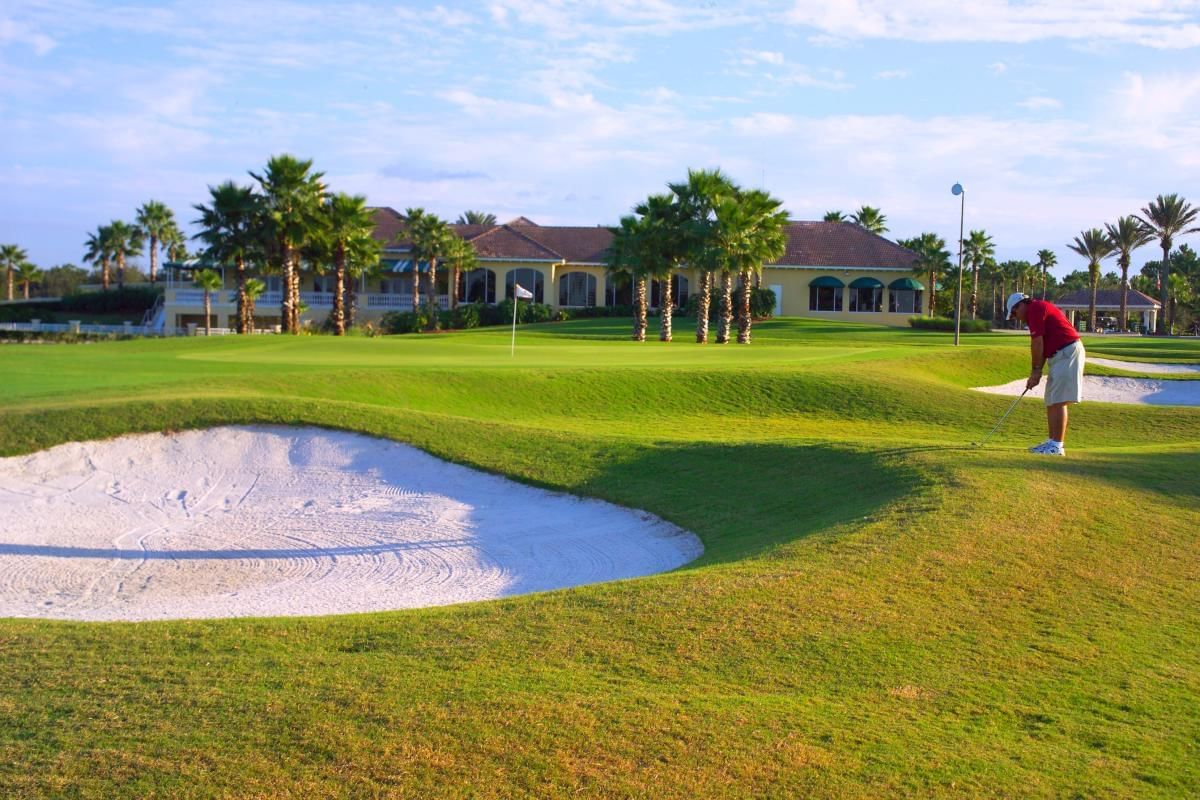 A golfer in a red shirt and white shorts is playing on a golf course with a sand trap in the foreground and a large building with palm trees in the background.