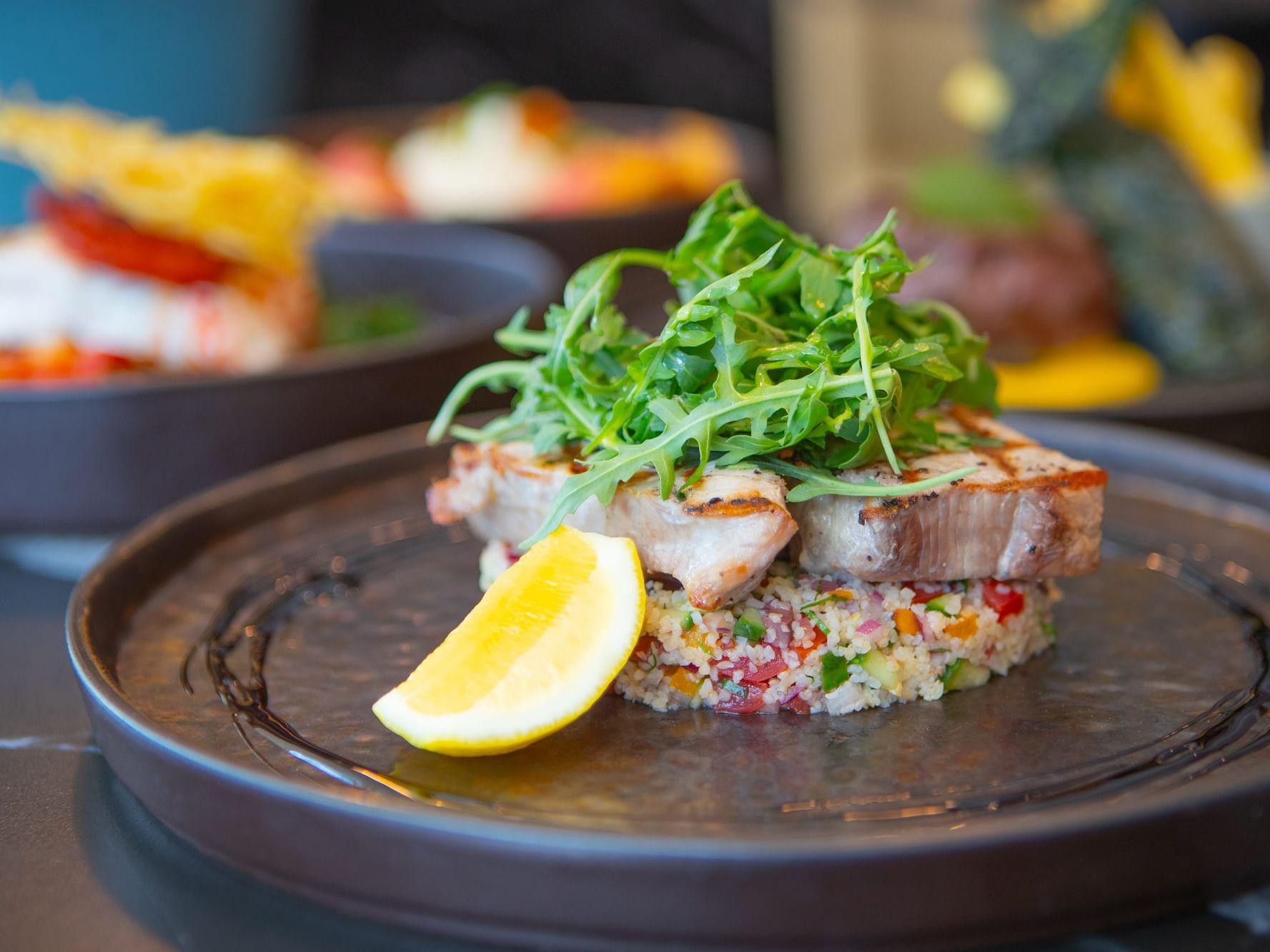 Plate of fish atop couscous with lemon wedge and arugula garnish, surrounded by other dishes.