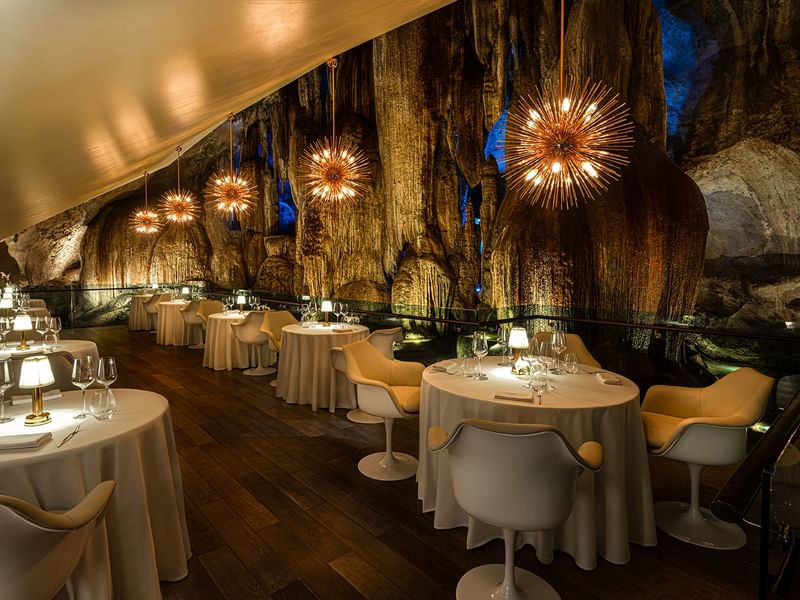 Elegant cave restaurant with round tables, white chairs, and lit starburst chandeliers hanging from the ceiling.