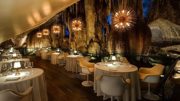 Elegant cave restaurant with round tables, white chairs, and lit starburst chandeliers hanging from the ceiling.