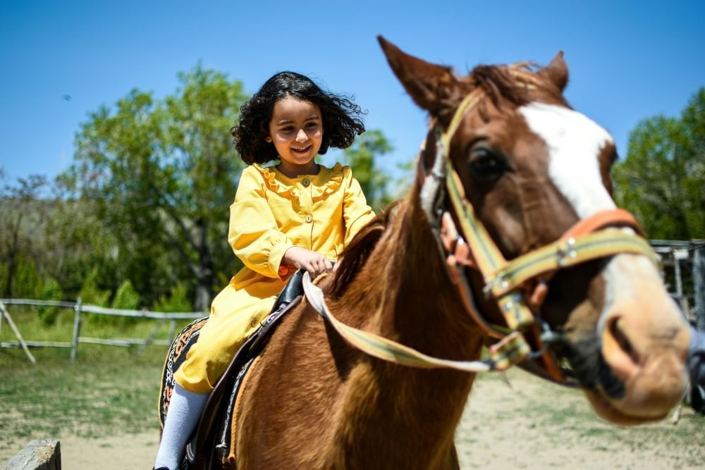 A young girl in a yellow dress smiling as she rides a horse with trees and a clear blue sky in the background.