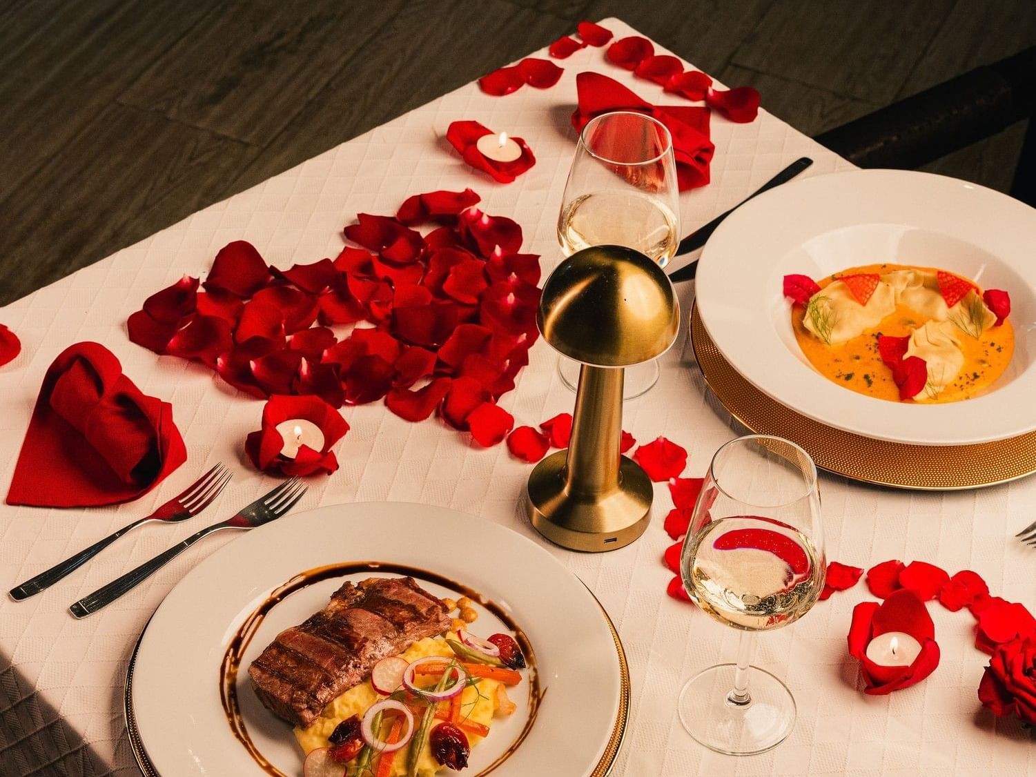 Intimate table for two with gourmet meals, wine glasses, scattered red rose petals, and glowing candles for a romantic dinner
