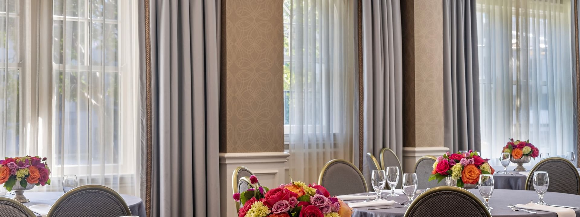 Round tables with floral centerpieces by grey curtains under soft window light in Oak Lawn/Terrace at Warwick Melrose Dallas