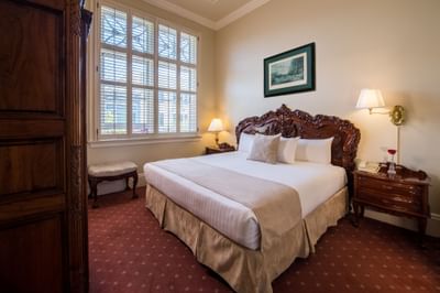 Bedroom with large bed, two nightstands, window with shutters, wooden door, and wall-mounted lamp and framed art.