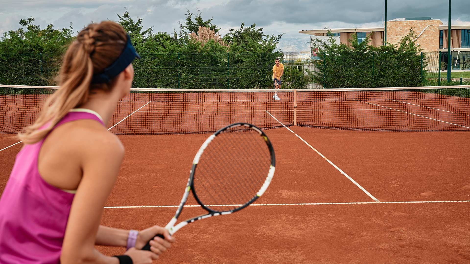 Žena s reketom igra tenis na terenu, muškarac je nasuprot.