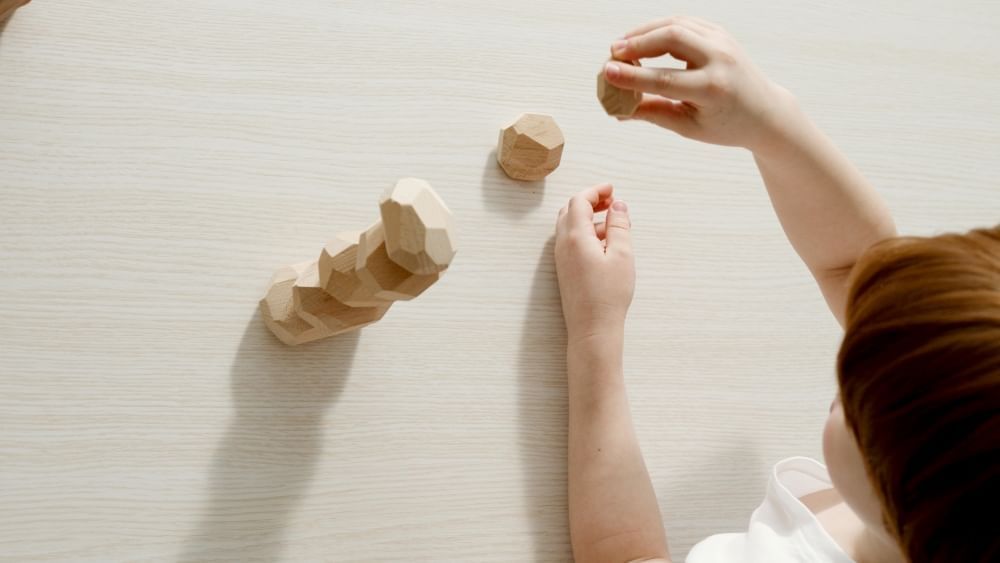 An overhead shot of a child stacking wooden geometrical blocks on a wooden table.