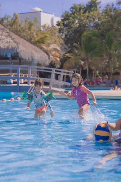 Two young girls wearing floaties splash and play in a sunny resort pool at Playa Blanca Beach Resort