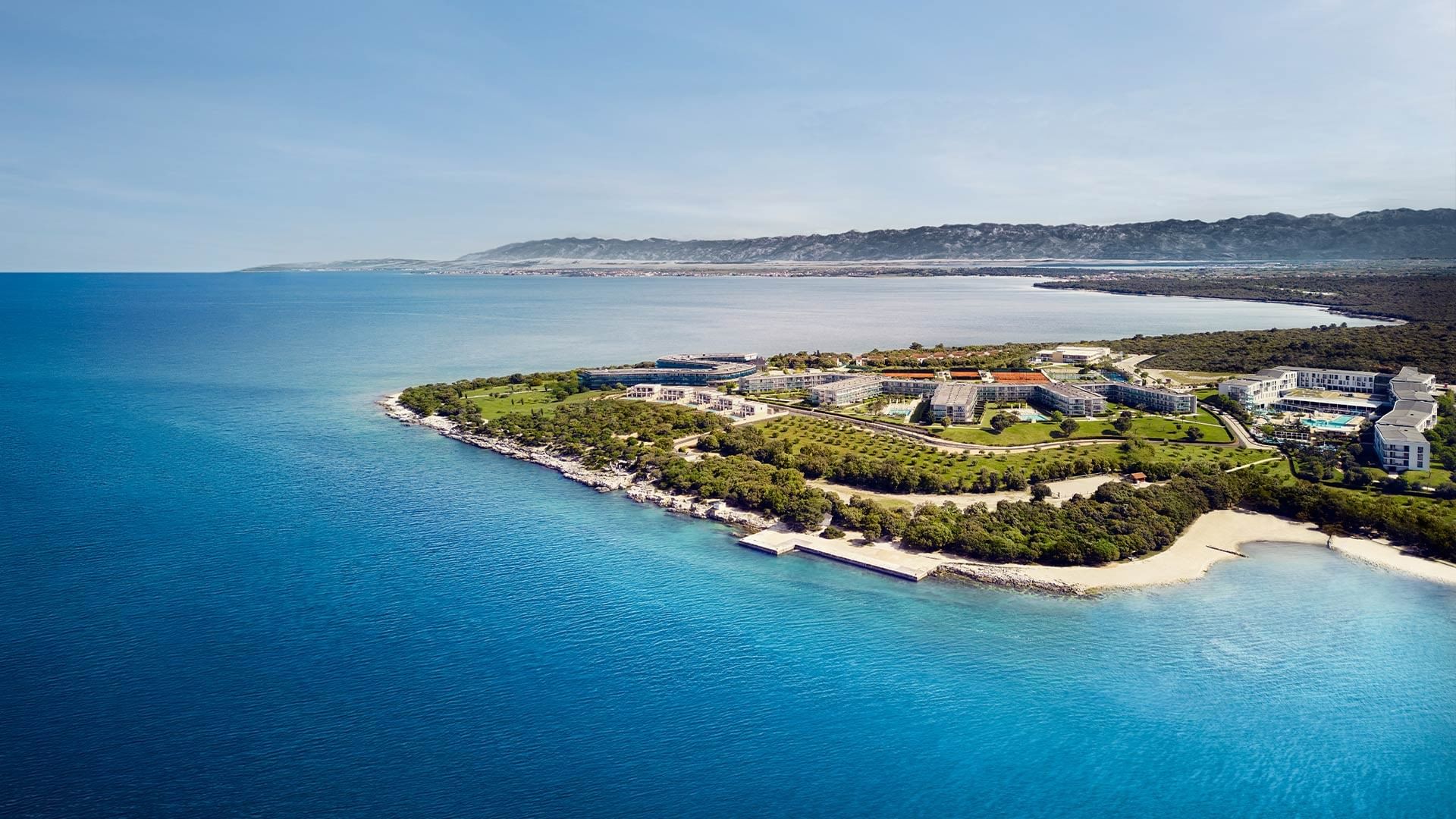Aerial view of a coastal resort with buildings and greenery surrounded by ocean at Falkensteiner Group