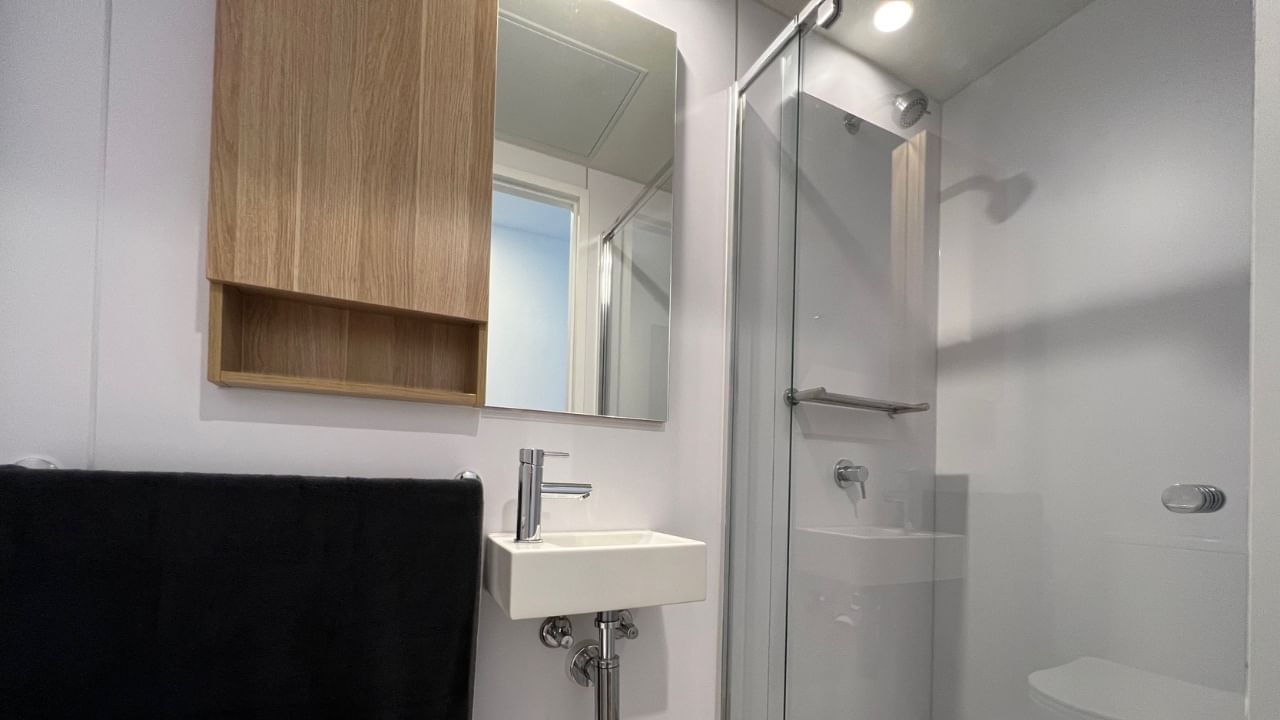 White bathroom with a wooden cabinet, mirror, glass shower enclosure, black towel, and white sink.