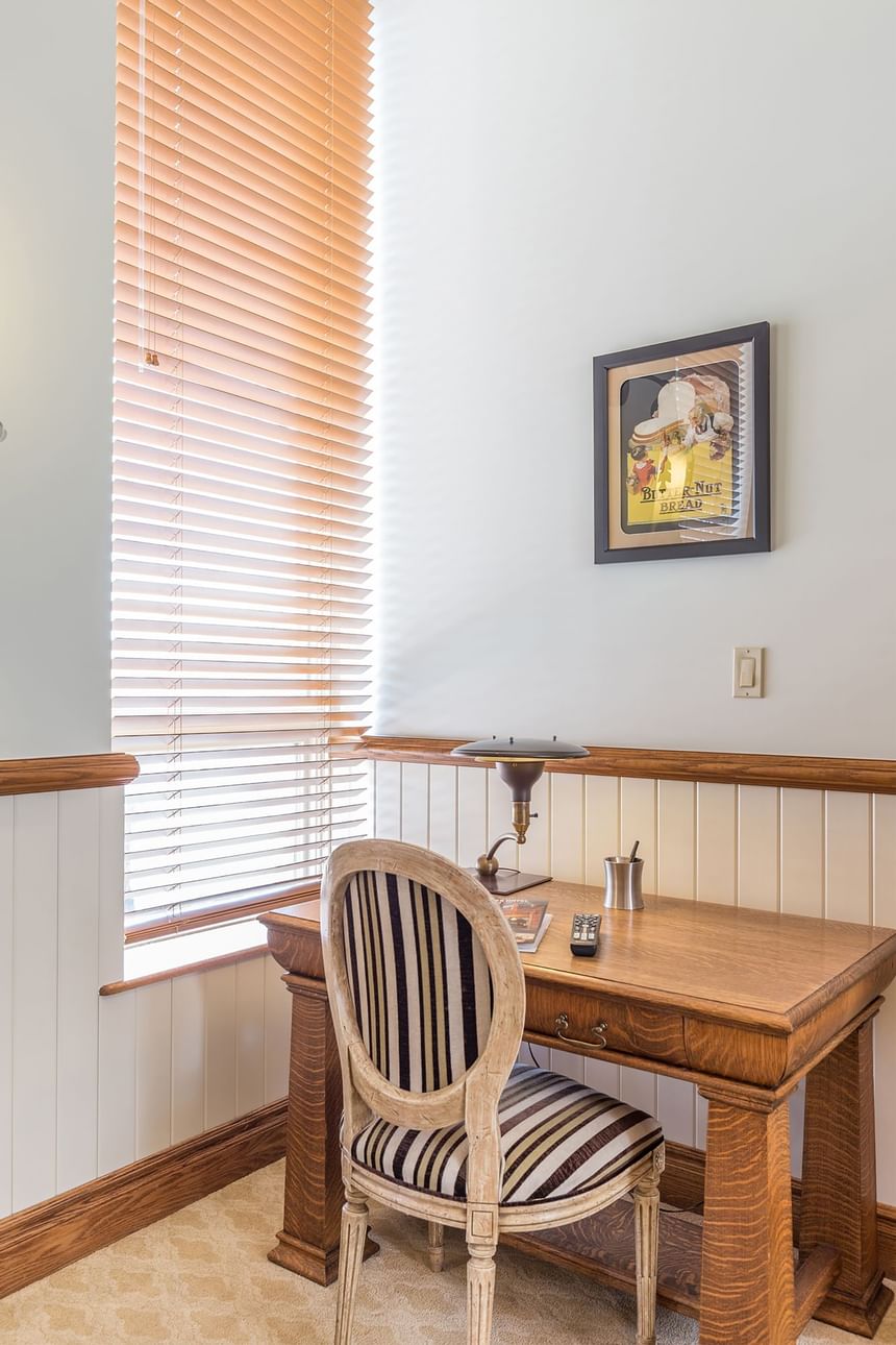 a chair faces a wooden desk with a desk lamp near a big window 