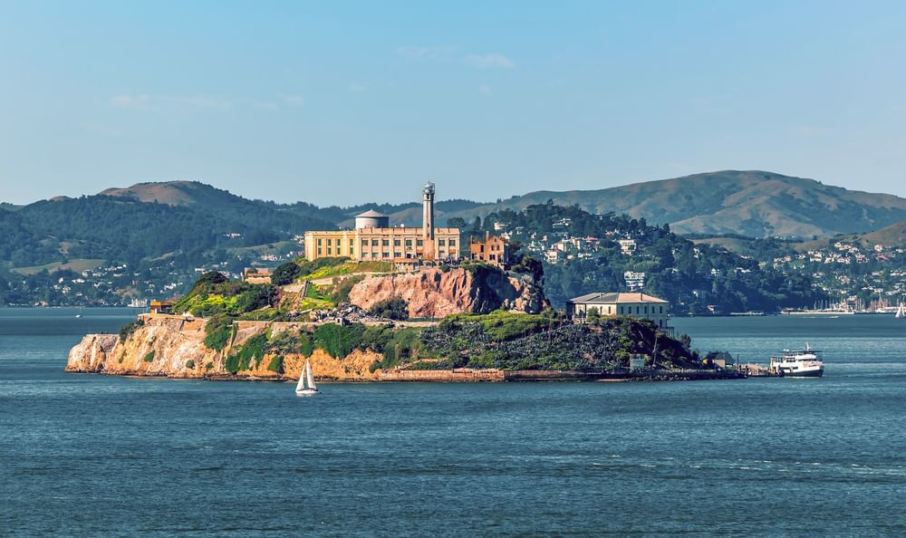 Alcatraz Island by a sailboat under a blue sky near the hilly coastline near Warwick San Francisco