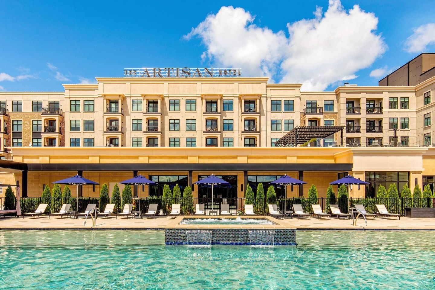 Artisan Hotel with pool, lounge chairs, and blue umbrellas under a blue sky with clouds.
