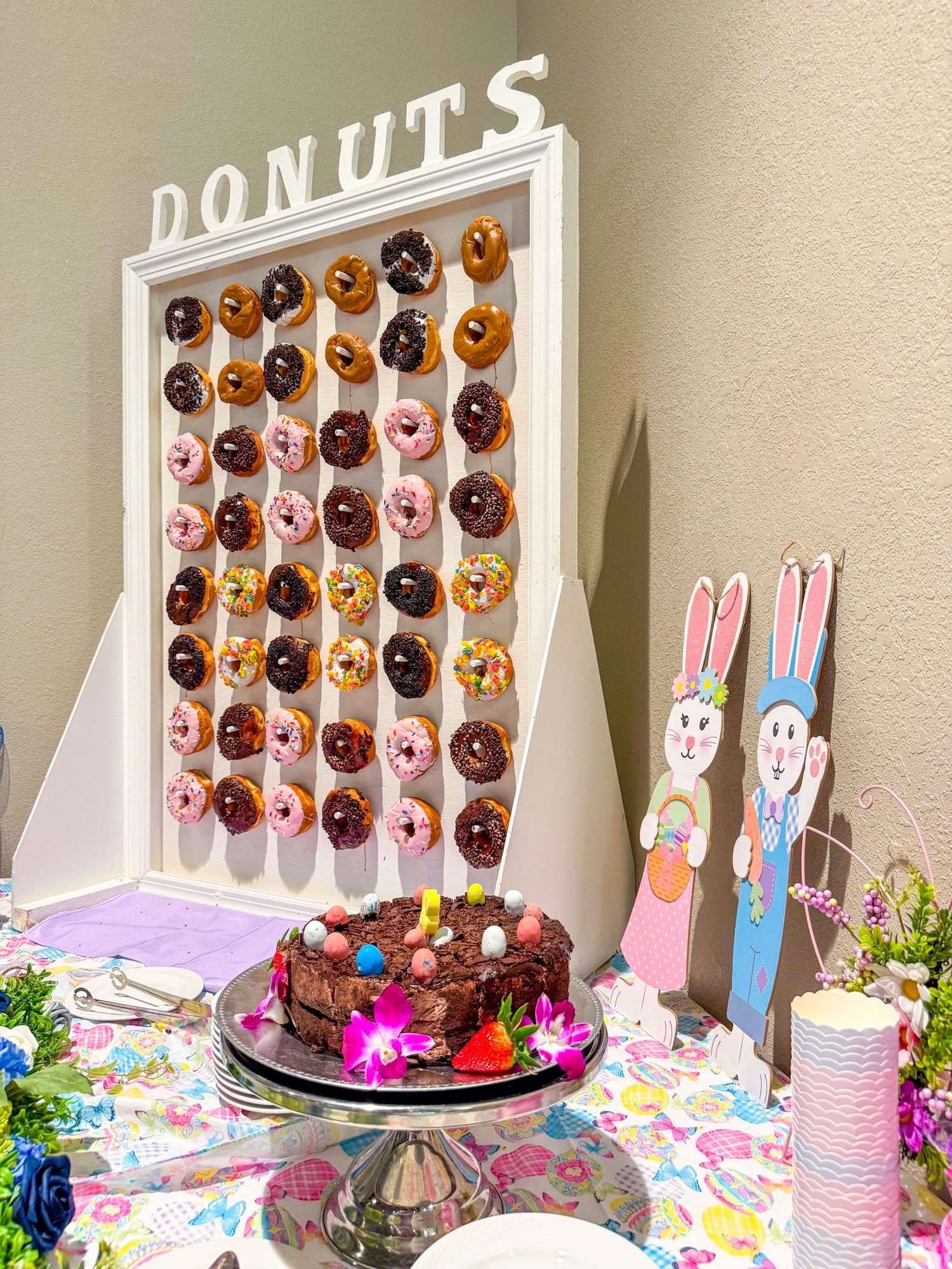 A large white board with pegs and hanging chocolate, pink, and sprinkled donuts, beside a cake stand with a decorated chocolate cake and two Easter Bunny decorations.  