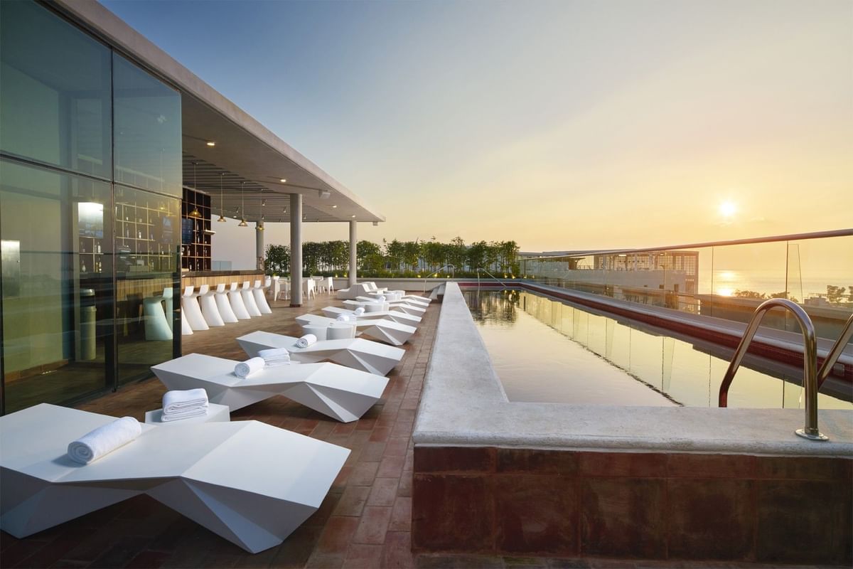 Terraza de piscina en la azotea con camastros blancos y bar con vista al atardecer en Fiesta Inn
