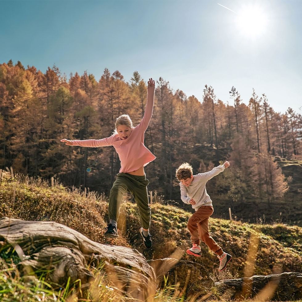 Due bambini saltano su un sentiero boschivo con il sole che brilla dietro di loro.