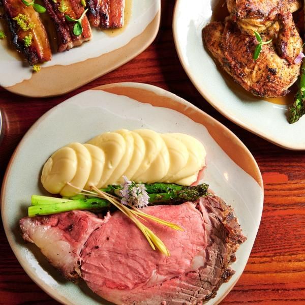 An overhead shot of three plates, one with roast chicken, one with candied bacon slabs, and one with a slice of prime rib, mashed potatoes, asparagus, and a floral garnish.