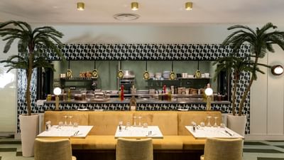 A modern restaurant interior with patterned walls, a kitchen counter, dining tables, and tropical plants.