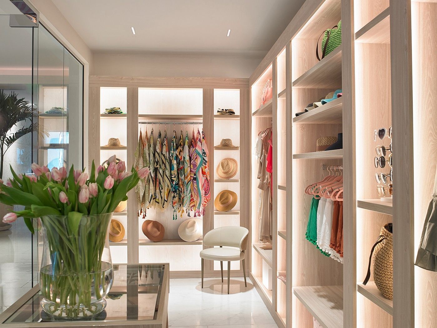 A large walk-in closet with light wood shelves and organizers, a chair, and a vase of pink tulips.