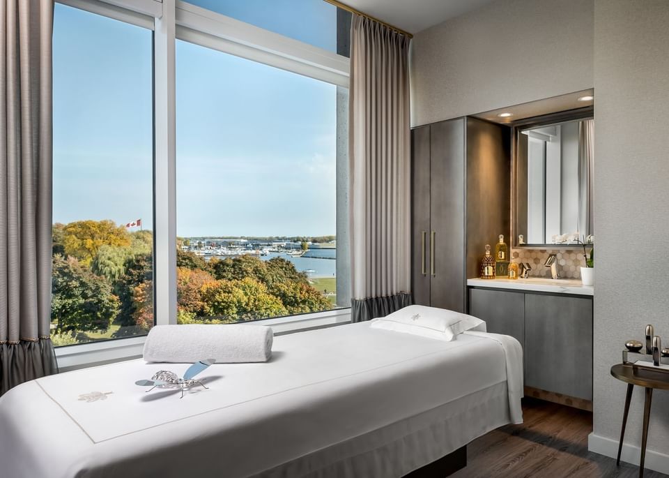 Quiet spa treatment room with a massage bed and vanity sink facing large window with scenic lake view at Hotel X Toronto