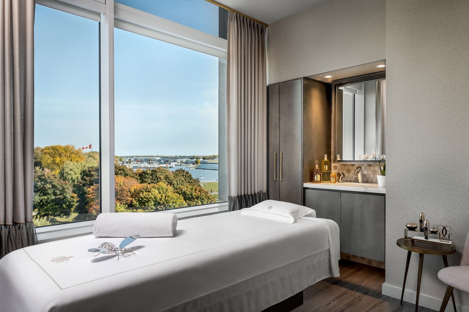 Quiet spa treatment room with a massage bed and vanity sink facing large window with scenic lake view at Hotel X Toronto