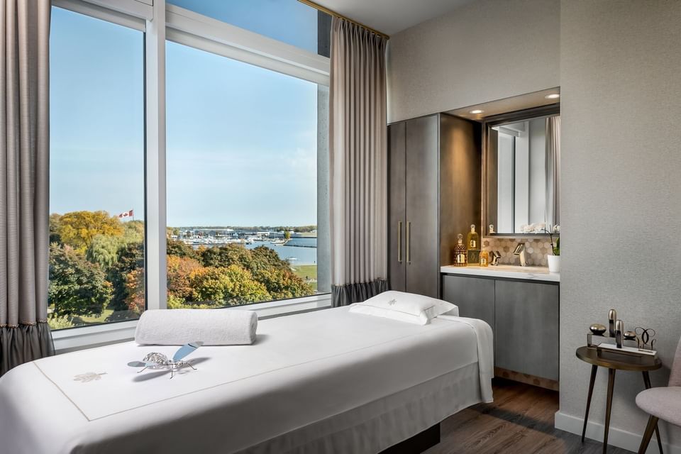 Quiet spa treatment room with a massage bed and vanity sink facing large window with scenic lake view at Hotel X Toronto