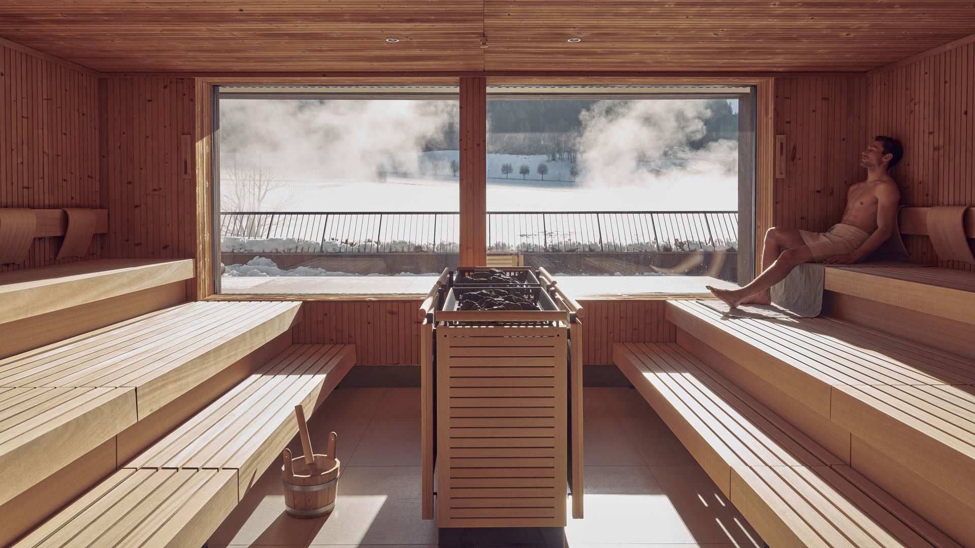 Man relaxing in a wooden sauna with a snow-covered view outside the window at Falkensteiner Hotel Kronplatz