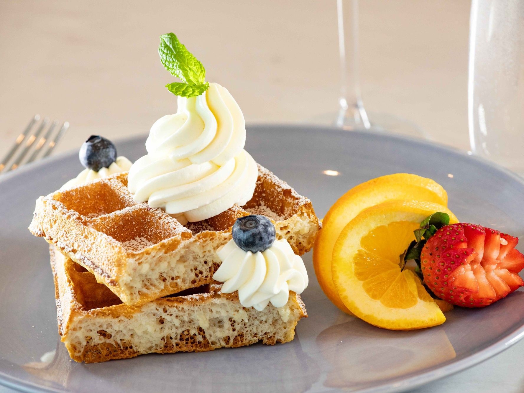 Golden waffles topped with whipped cream, blueberries, and mint, served with fresh orange slices and strawberries.