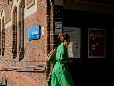 Woman in flowing green dress walking briskly past historic brick train station entrance with arched windows.