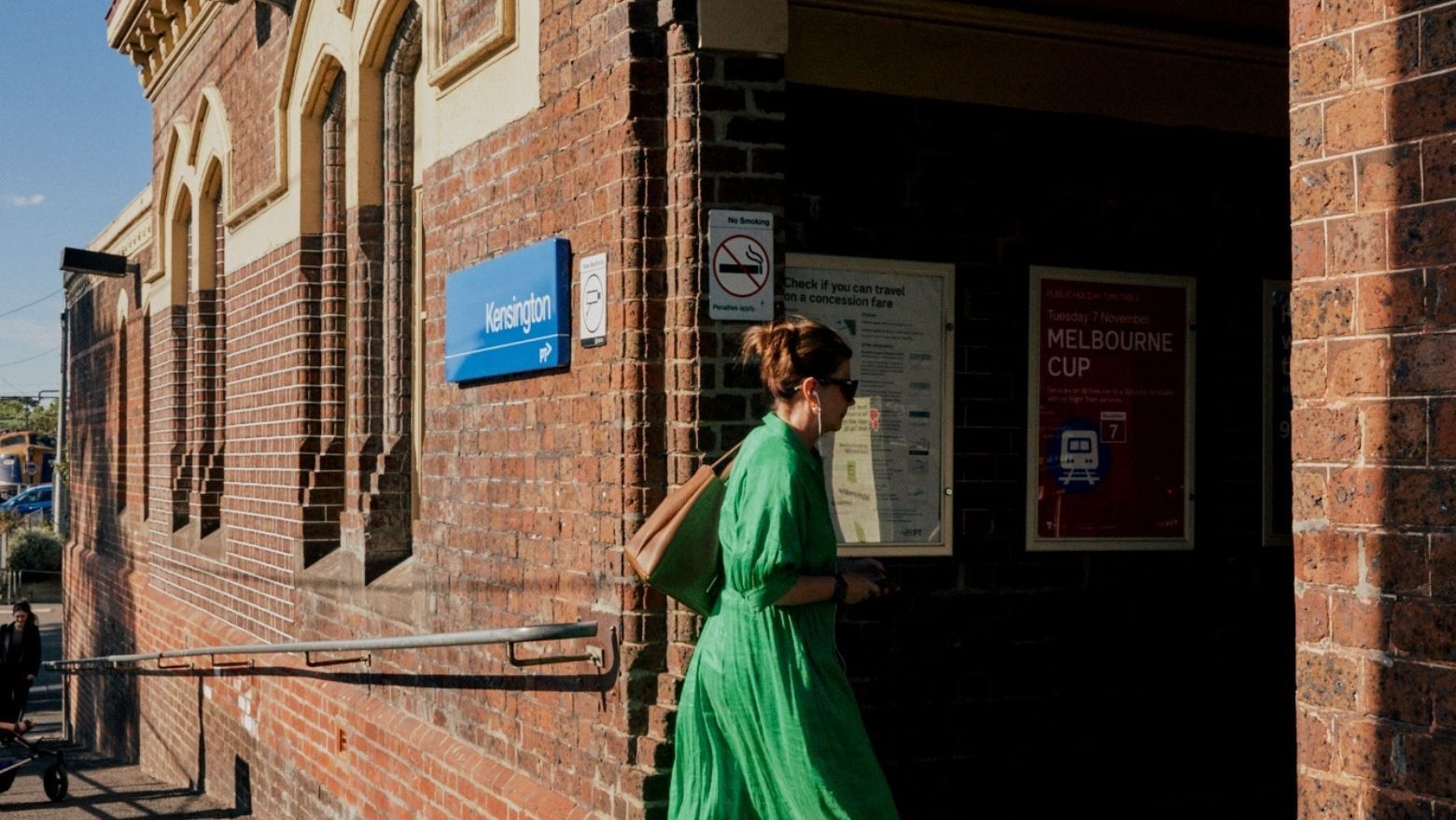 Woman in flowing green dress walking briskly past historic brick train station entrance with arched windows.