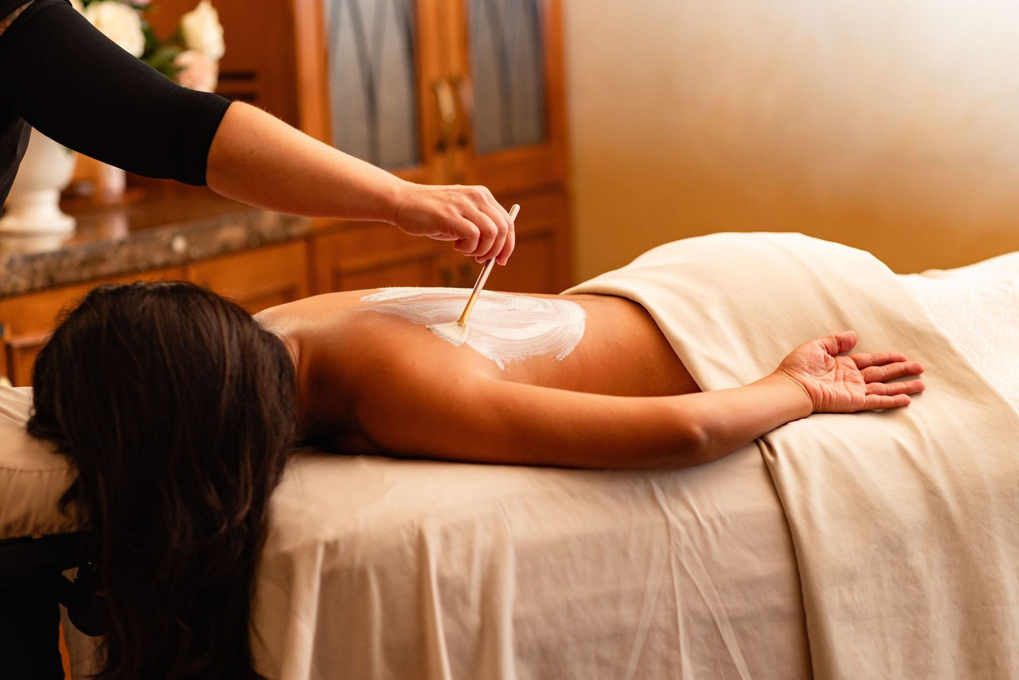 Spa professional using a brush to apply a white mud mask to a client's back during a treatment at Kelly's Spa