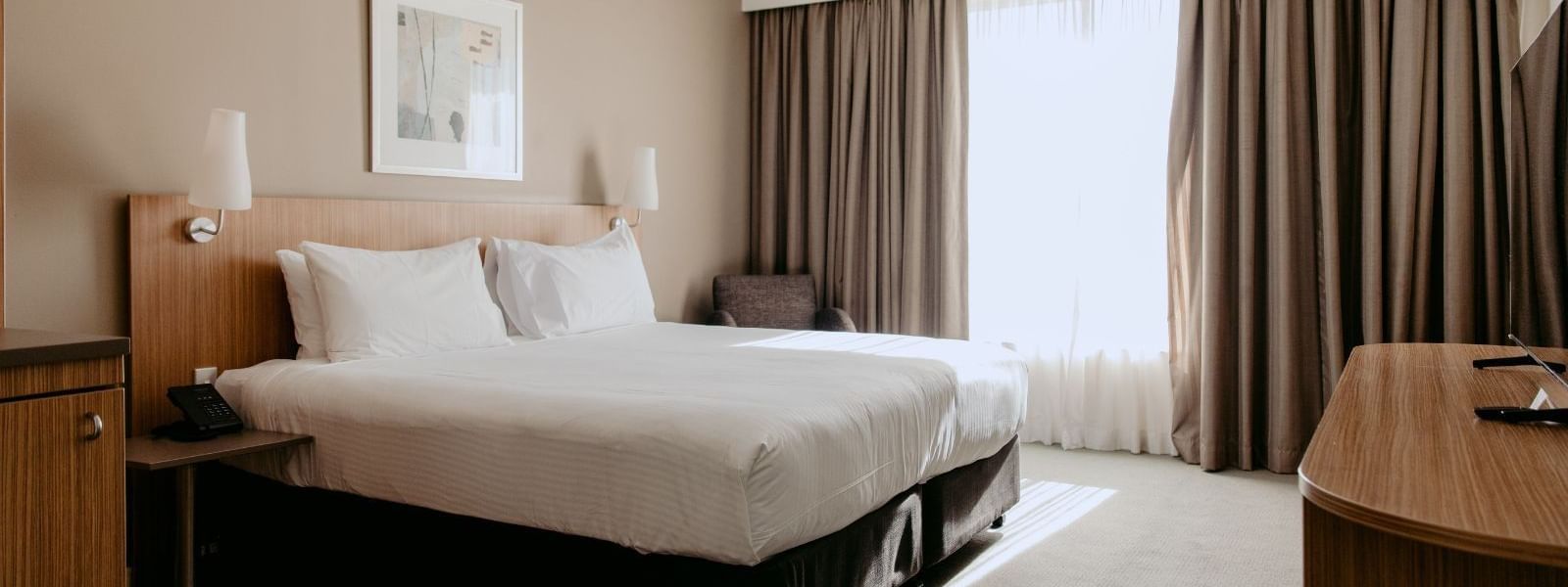 A hotel room with a bed, two lamps, a desk, a chair, and a large window.