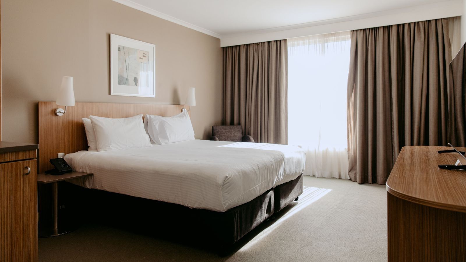 A hotel room with a bed, two lamps, a desk, a chair, and a large window.