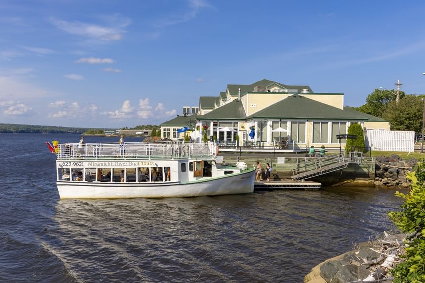 Tour boat docked by a wooden ramp under a sunny sky, with a yellow waterfront resort at Rodd Miramichi River