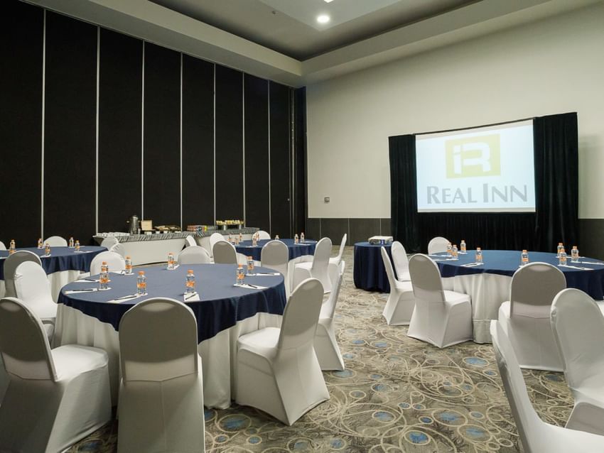 Banquet set-up with blue linens, a projector screen, and a refreshment corner in the Verona Venue at Real Inn Tijuana