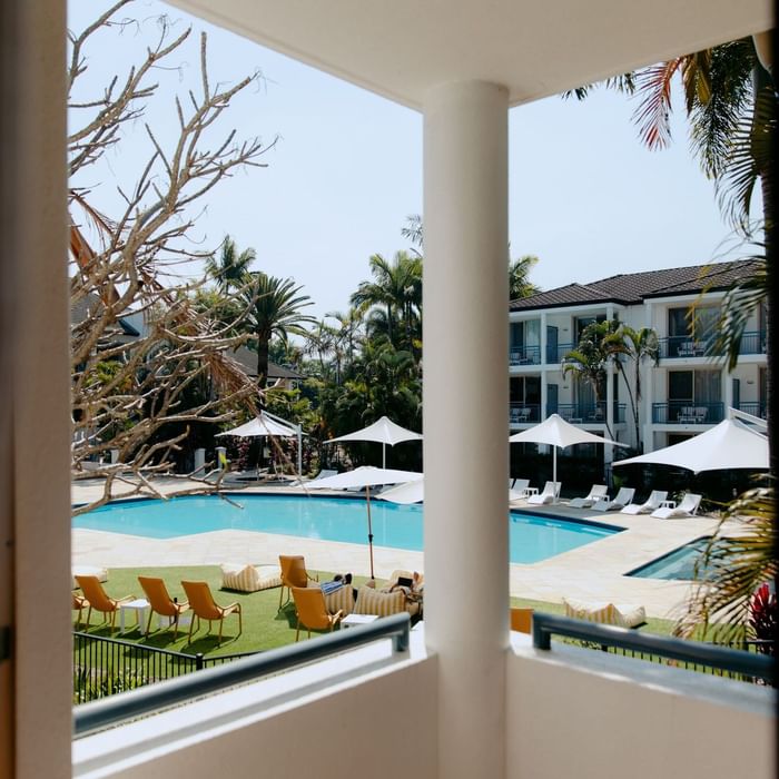 View from a window showing a resort with a pool, lounge chairs, umbrellas, and buildings.