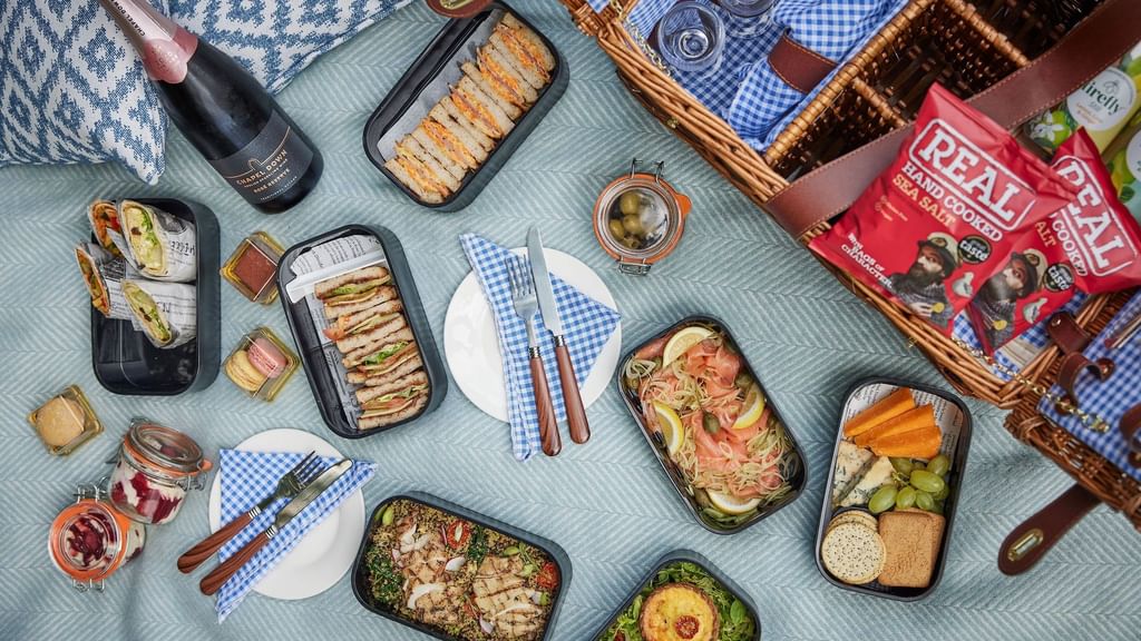 Picnic spread on a blanket with sandwiches, fruits, cheese, crackers, and a bottle of wine.