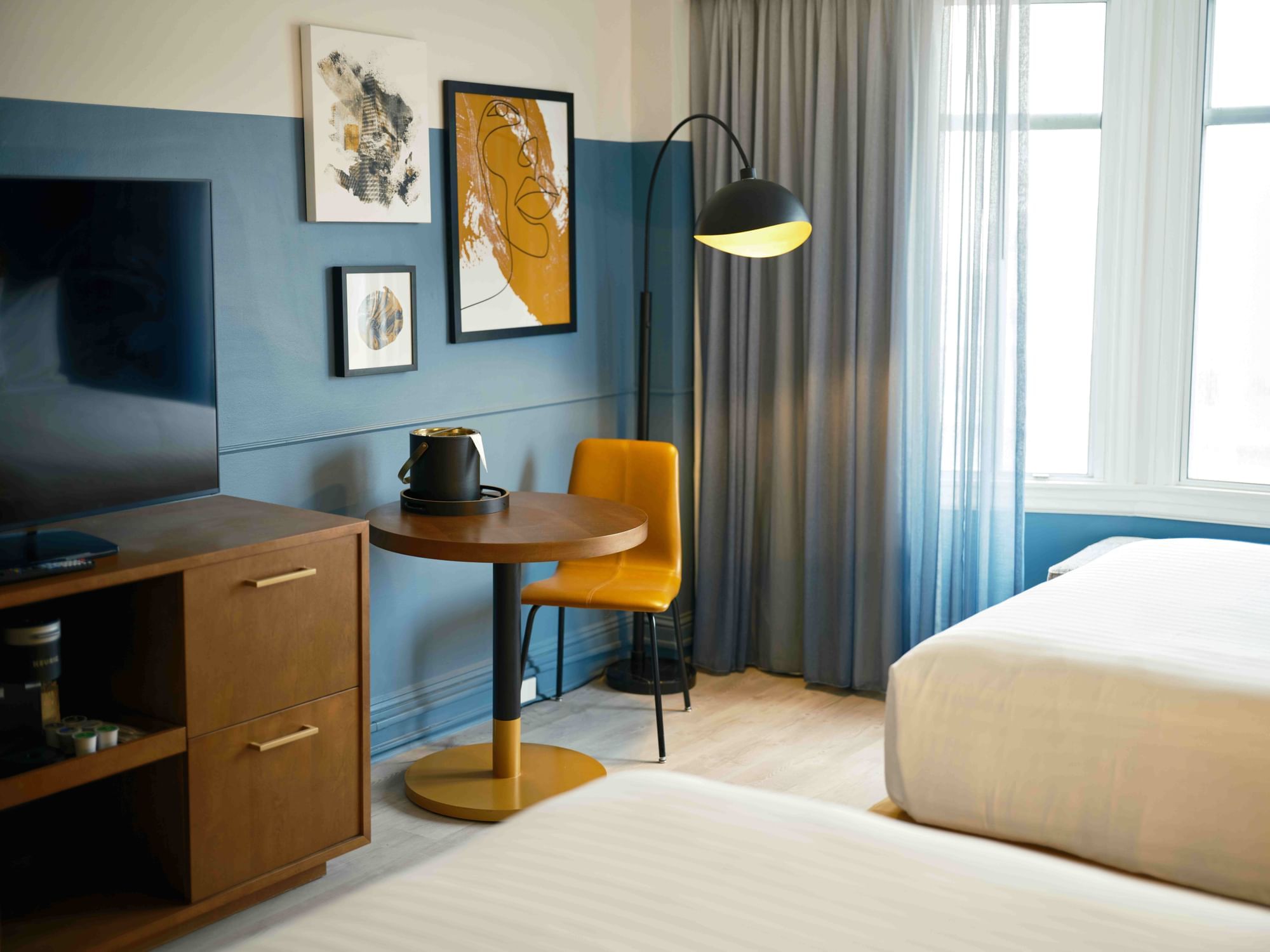 A hotel room with a bed, desk, TV, and artwork on the wall.
