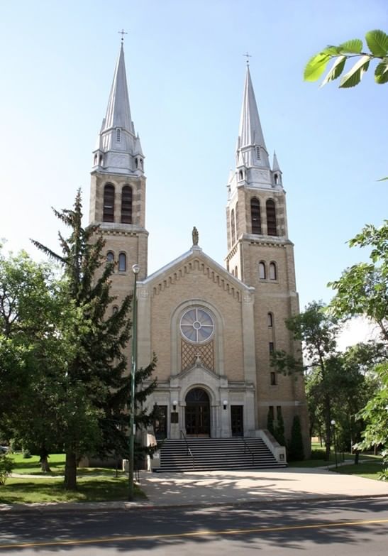 Photograph of a large church building with two tall, pointed steeples and a central circular stained glass window above the main entrance. Surrounded by trees and greenery, the structure features light-colored brickwork and symmetrical architectural details, with steps leading up to double doors.