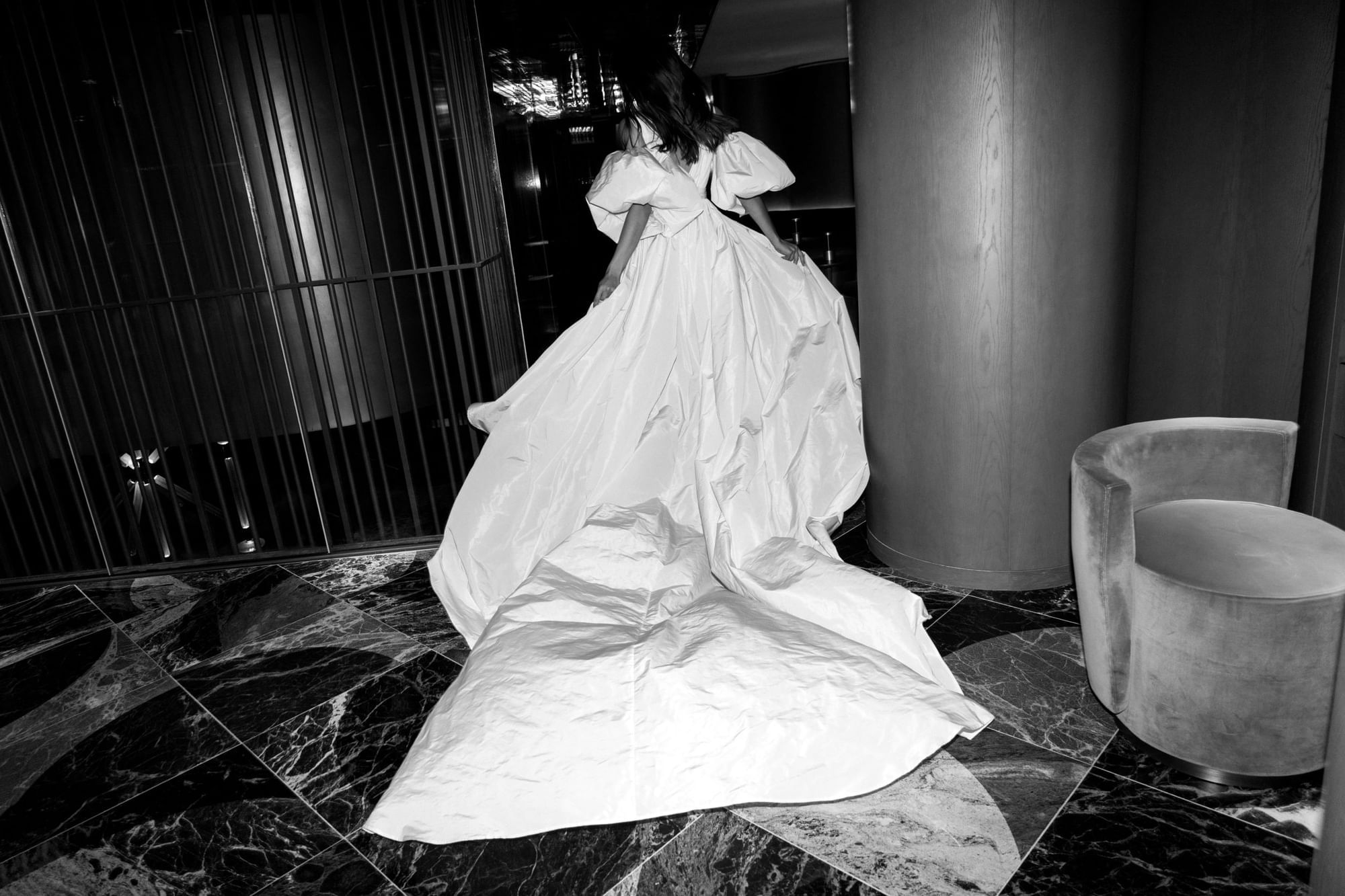 Black & white shot from behind of a woman in a white Olive Dress flowing across a marble floor at The Londoner Hotel