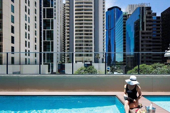 Woman in a sun hat sits by the hotel pool with champagne, enjoying the view of the city high-rises at The Sebel Brisbane
