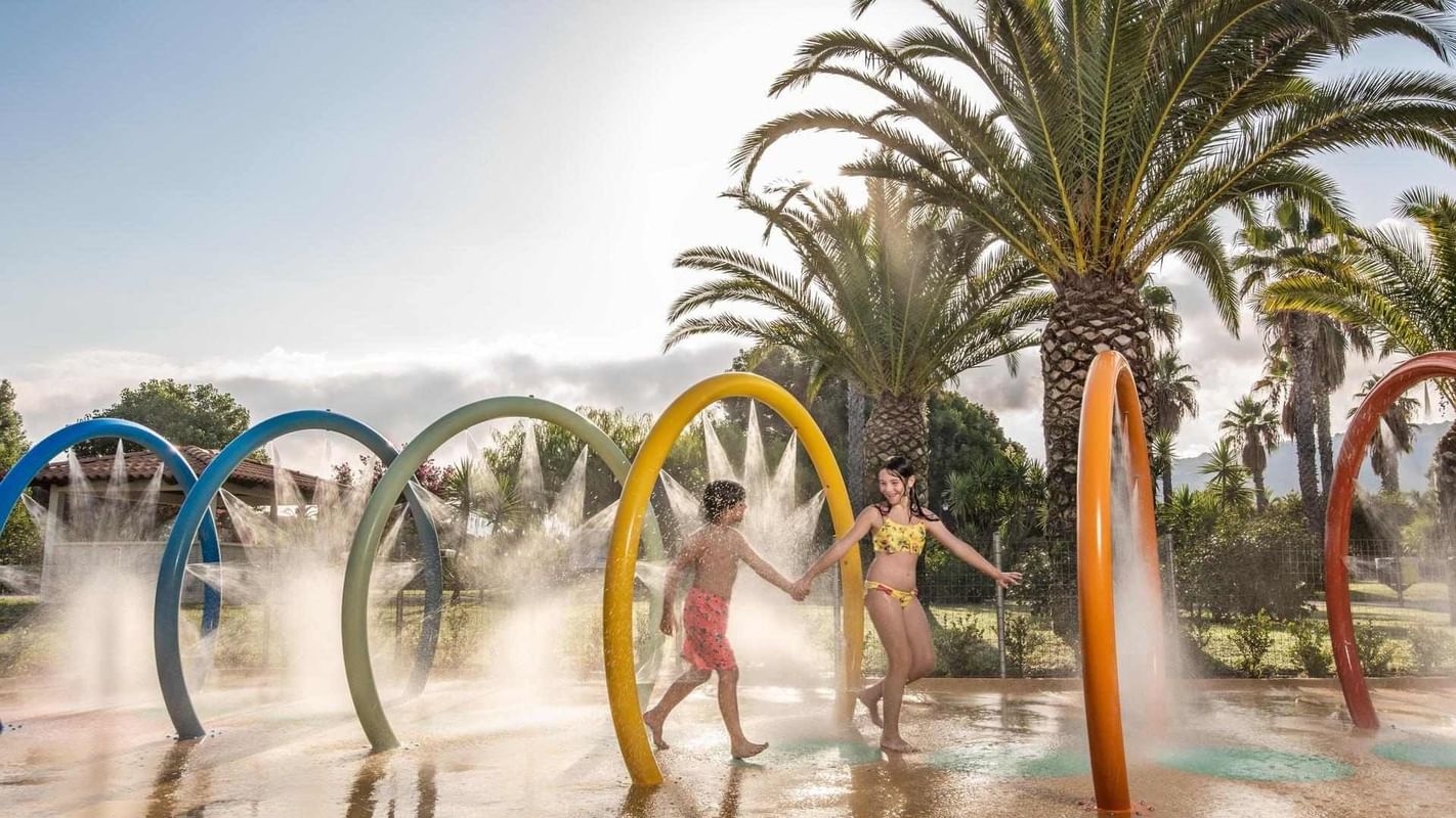 Bambino e ragazza che giocano sotto le fontane ad arco in un parco acquatico.