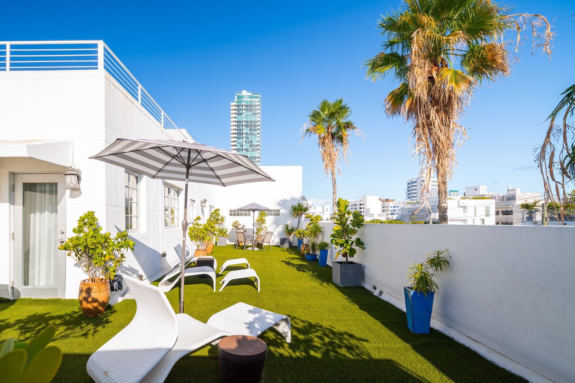A rooftop lounge with artificial turf, white lounge chairs, umbrellas, and potted plants overlooking the city.